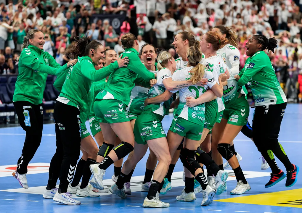 A group of athletes in green and white uniforms embrace and celebrate on a court, with a cheering crowd in the background.