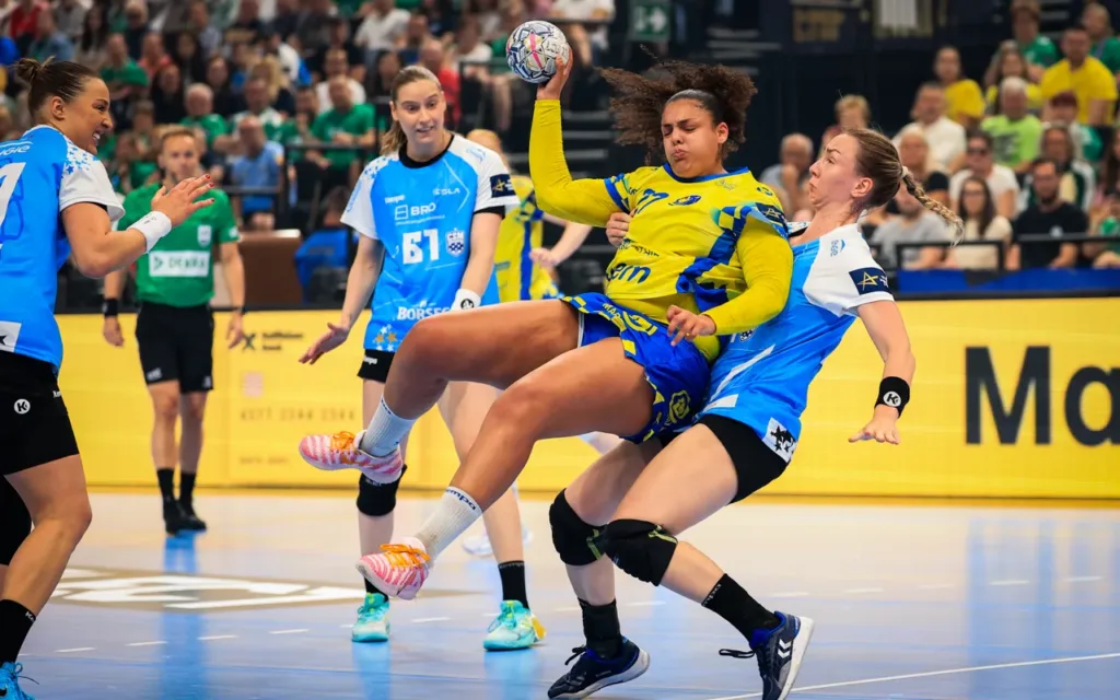 Bojana, Đina i Tanja bez finala: Mec i nesavladiva Bundsen ugasili snove Bukurešta 1 A player in a yellow and blue uniform leaps to pass the ball while being closely defended by an opponent in a blue jersey during an indoor handball match.